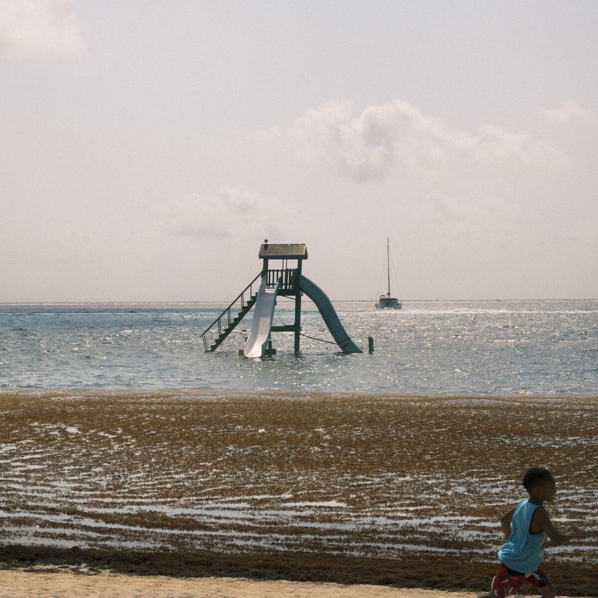 A photo of the horizon, water, a slide, a boat, and a kid running.